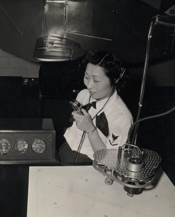 A woman in a military uniform wearing headphones sits while looking at an instrument.