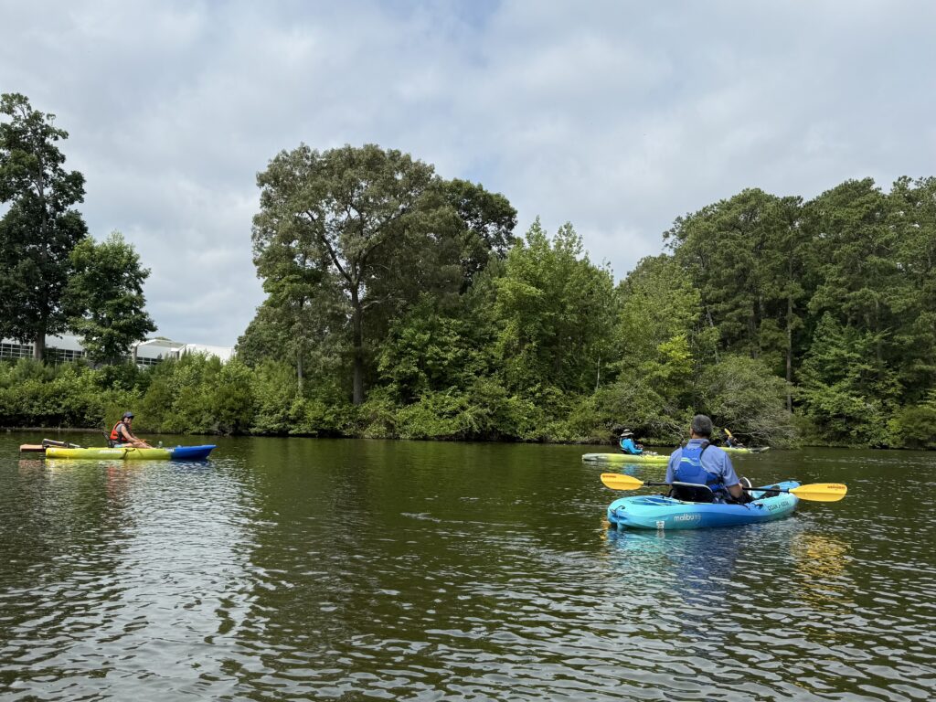 Three adult kayakers talking while floating on Mariners' Lake. Along the shore you can see The Mariners' Museum and trees.