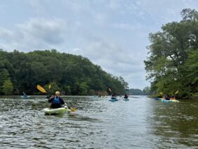 Adults and children kayaking on Mariners' Lake with lifejackets on. The sky is blue and the along the shore are trees.