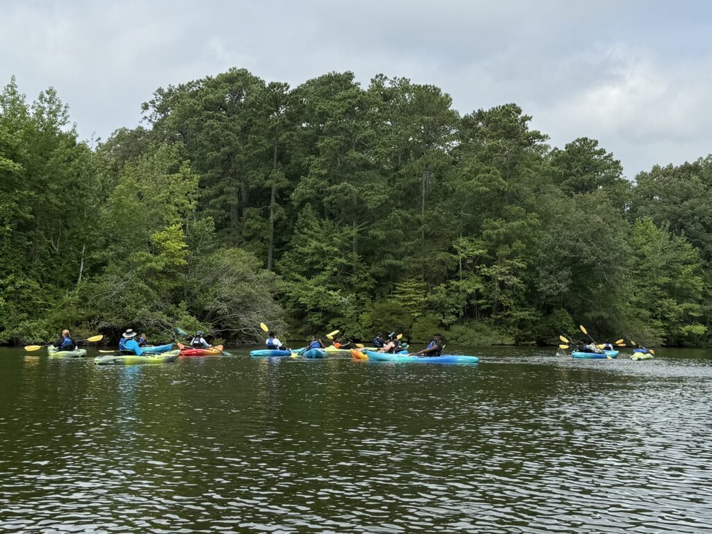 A group of children and adult kayakers floating and paddling around Mariners' Lake. Trees are in the background.