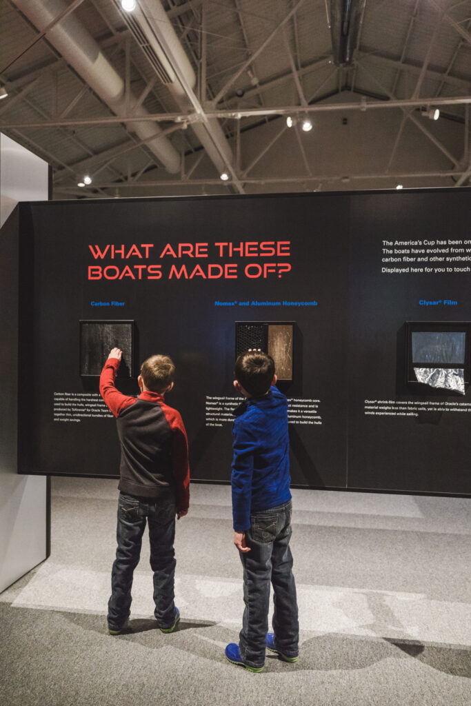 Two children in the Speed and Innovation in The America's Cup feeling the "What are these boats made of?" samples.