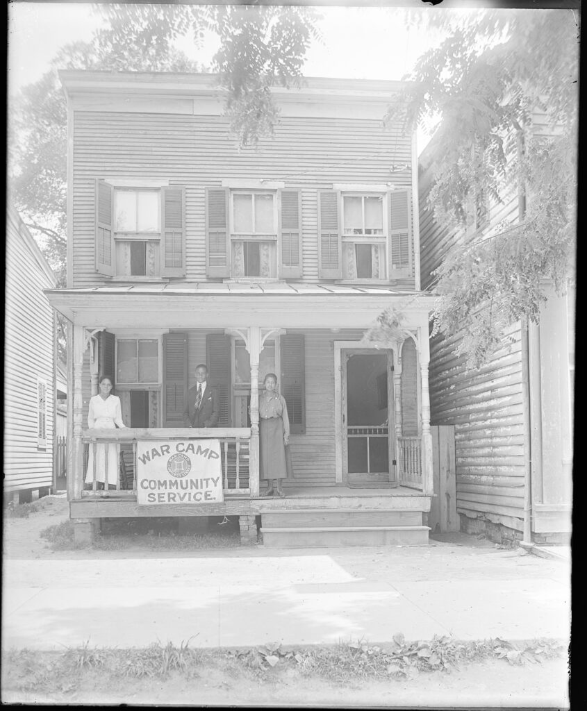 A black and white photo of 2 women and 1 man on a porch with the sign: War Camp Community Service