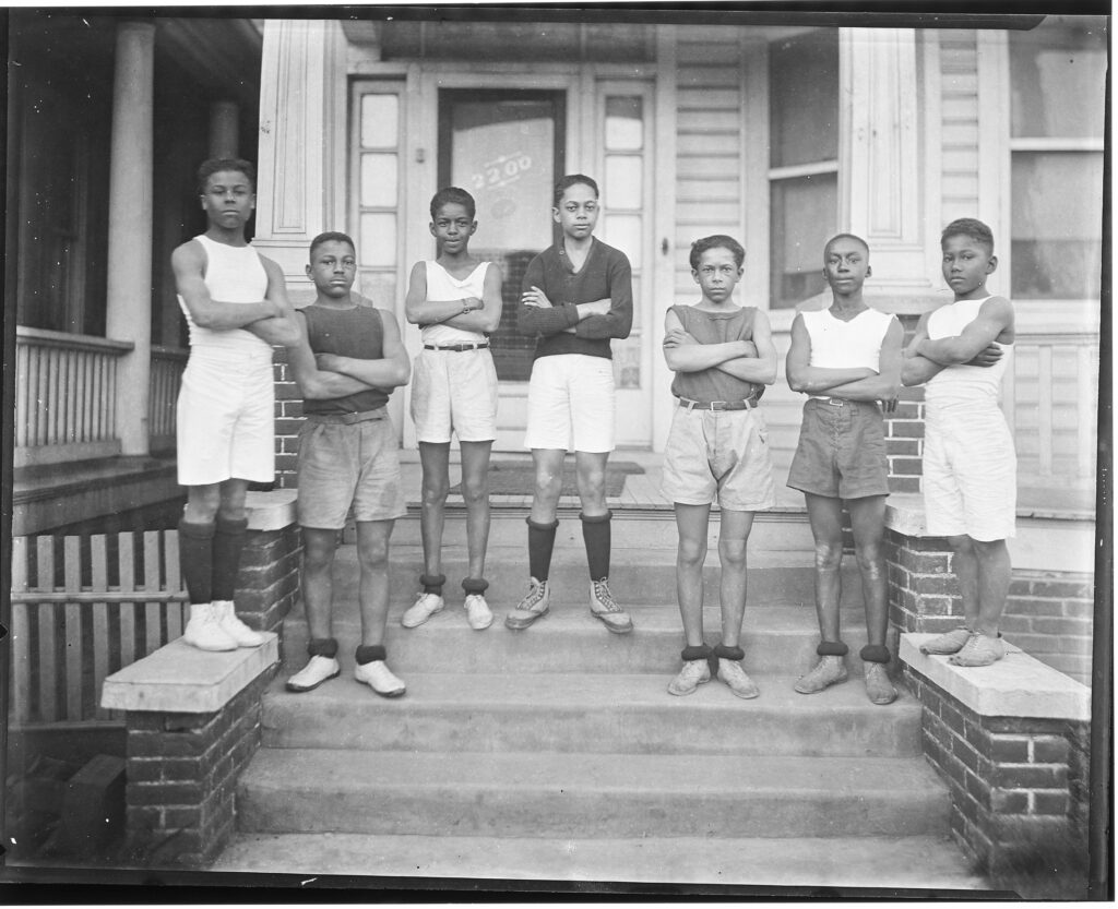 A black and white phot of several young boys on a stoop with their arms crossed.
