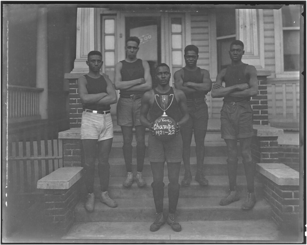 Black and white phot of 5 young men on a stoop. One is holding a trophy with a basketball attached.